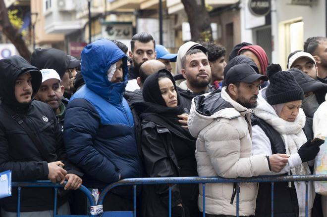 Grupo de personas con abrigos y ropa de abrigo, incluyendo mujeres con hiyab, congregadas detrás de una valla azul en una calle urbana con tiendas y bajo un cielo nublado.